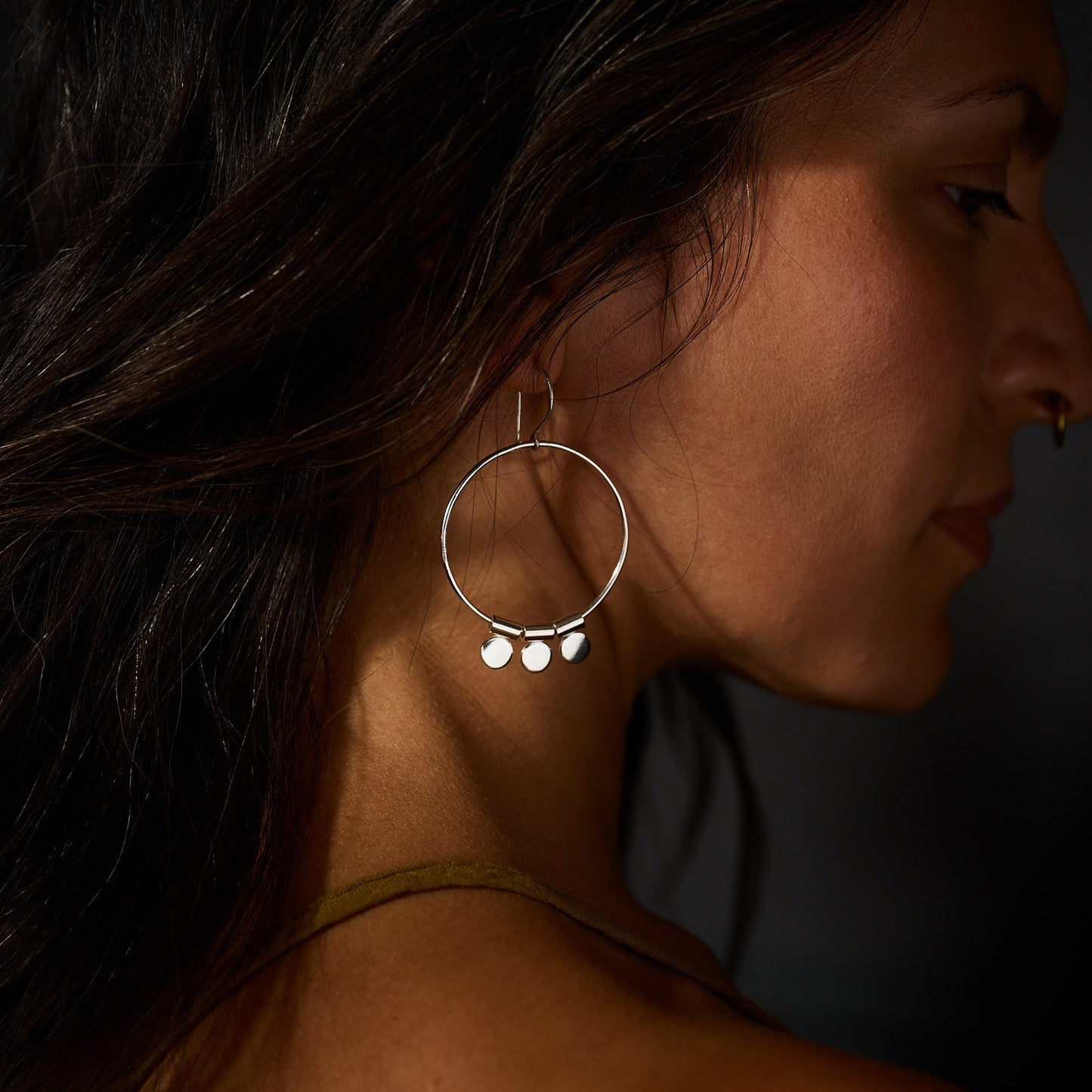 Close-up of a woman wearing a silver hoop earring with dangling charms against a dark background.