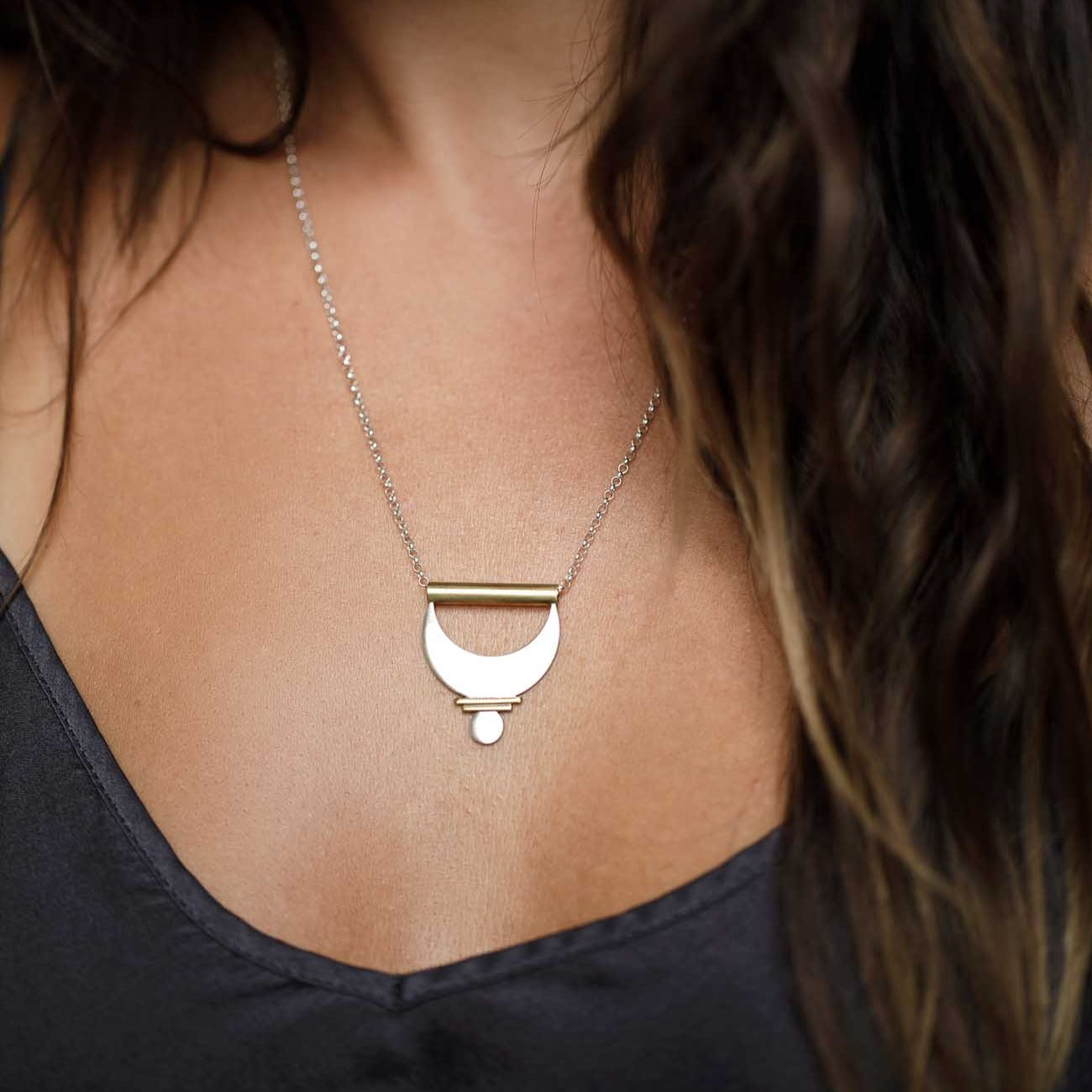 Wearing the Large Prana Pendant. Slivers of brass sun ray elements in between a large silver crescent moon and a small silver circle shape.
