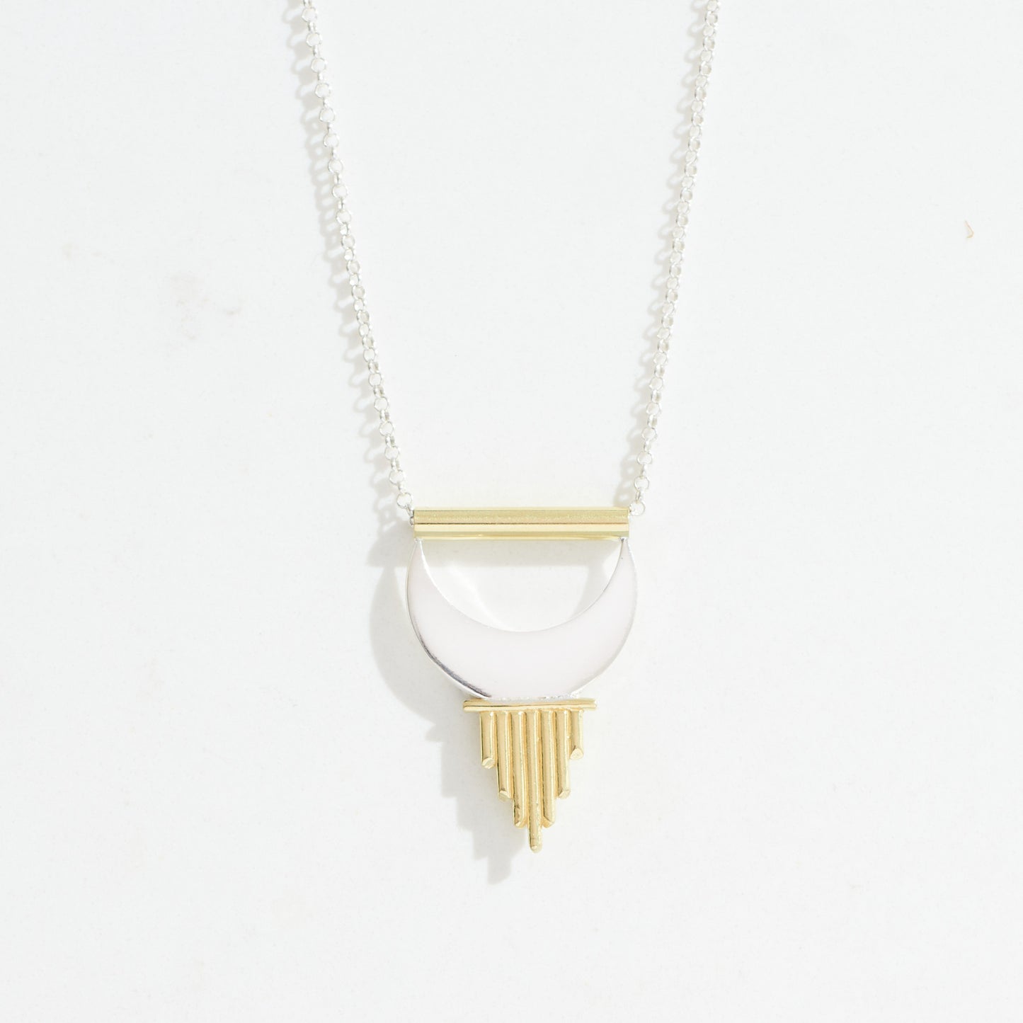 A necklace with a large pendant featuring a geometric design that includes a silver crescent moon shape with terminating in gold sun rays suspended from a gold tube that a silver chain runs through. 