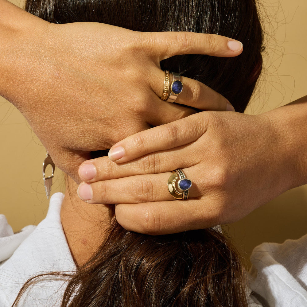 
                      
                        Close-up of hands with gold rings featuring blue stones against a beige background
                      
                    