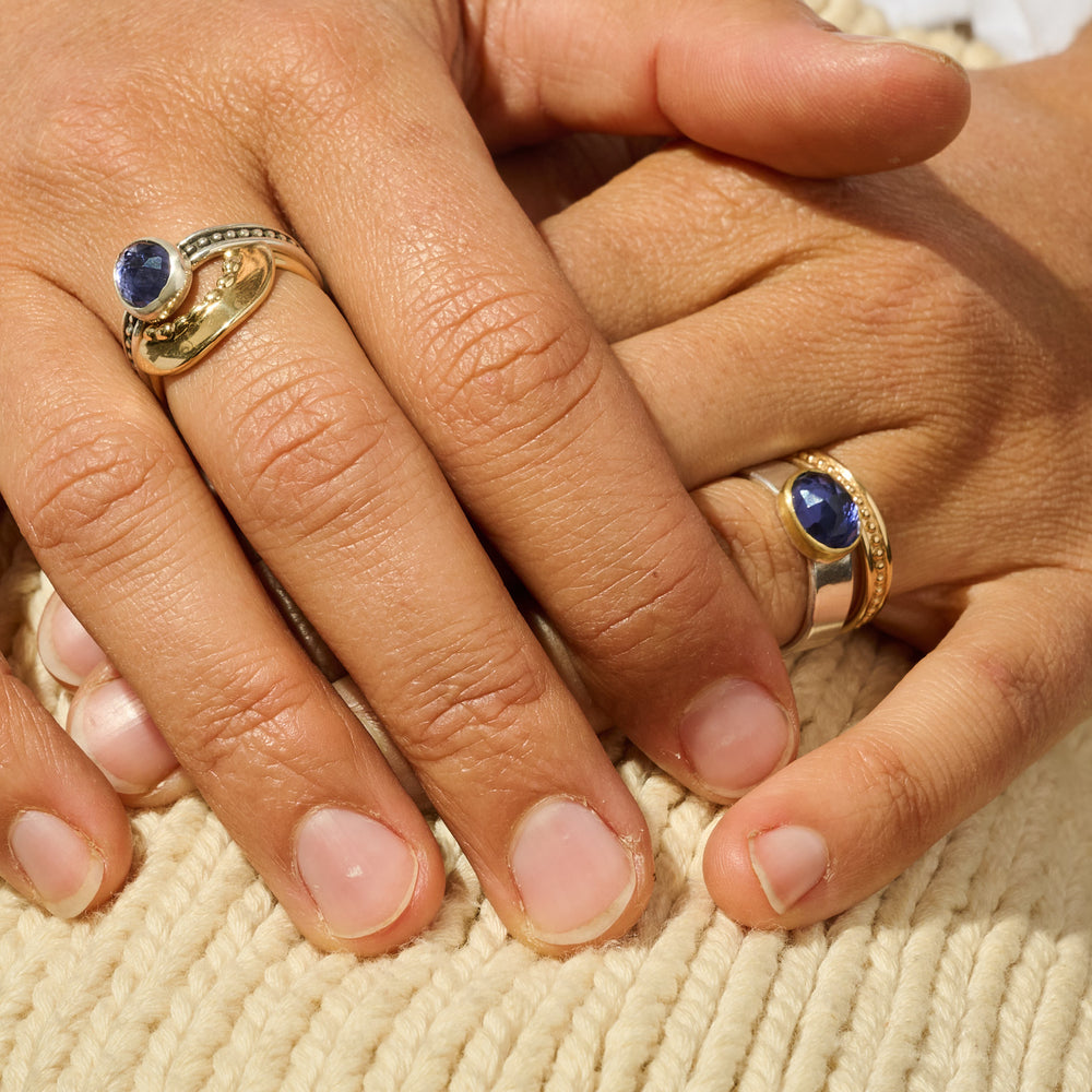 
                      
                        Close-up of two hands wearing gold rings with blue gemstones on a textured beige surface.
                      
                    