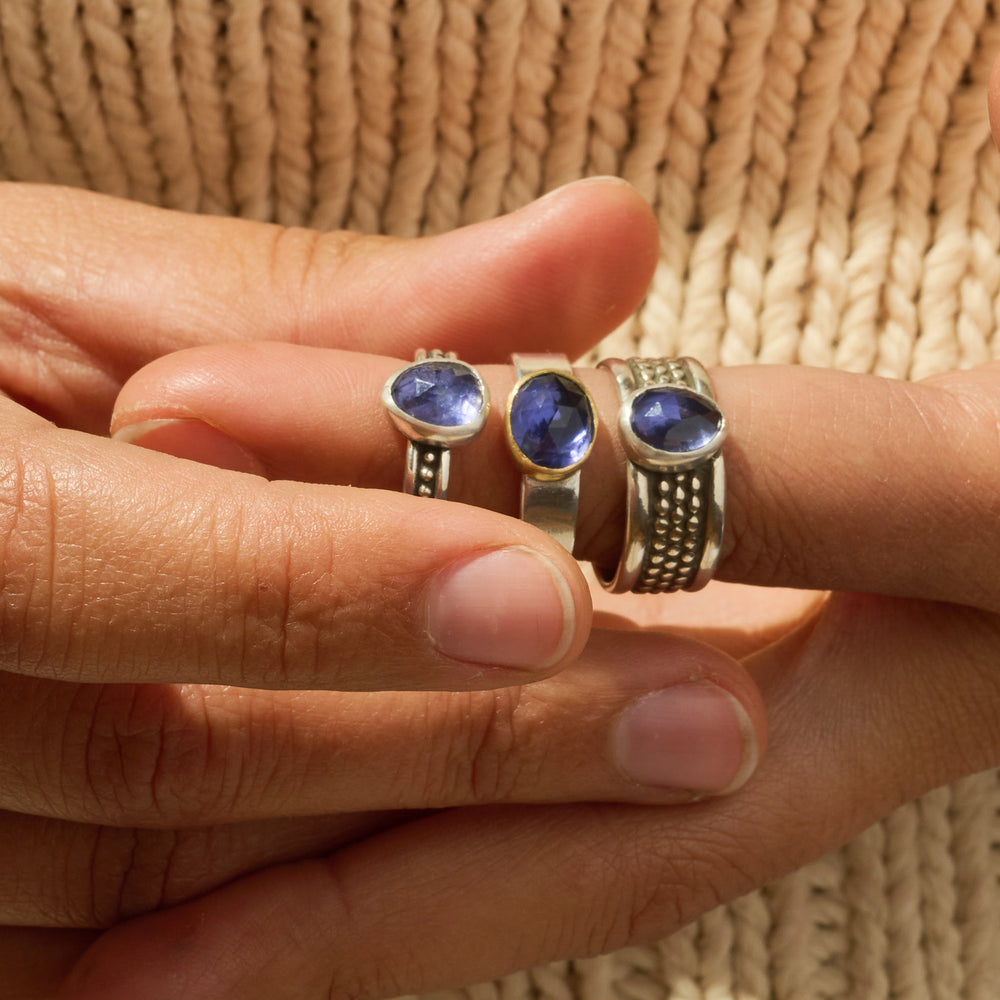 
                      
                        Three rings with blue stones on a hand against a textured beige background
                      
                    