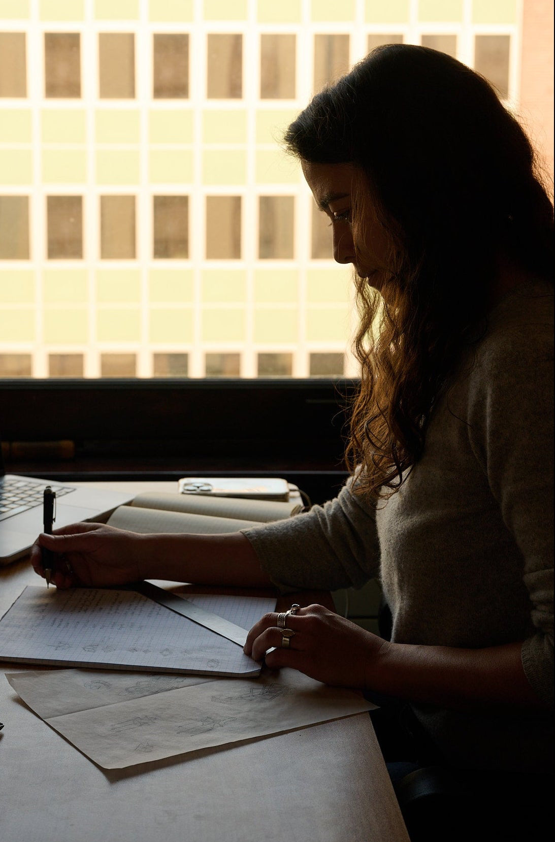 Woman working at a desk with a laptop and glass of wine, silhouetted against a window.