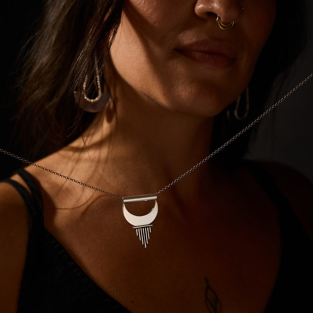 
                      
                        Woman wearing a necklace with a unique pendant against a dark background
                      
                    