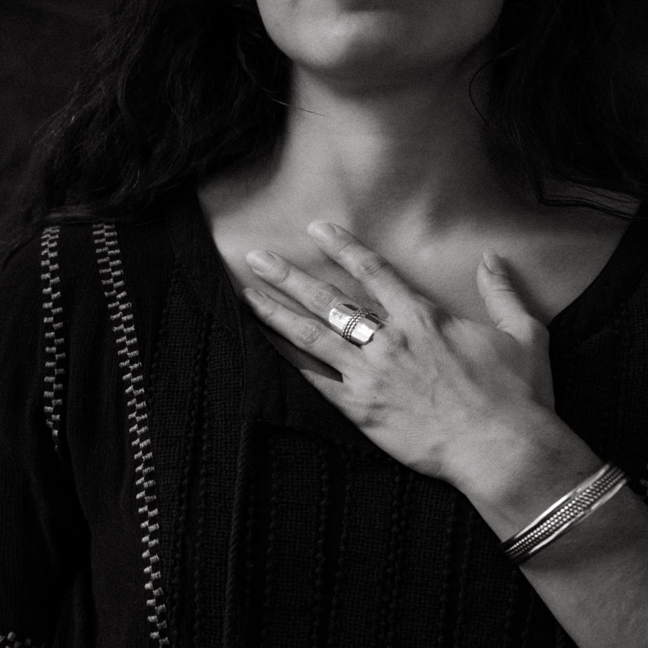 Person wearing a black garment, hand resting on chest, wearing a bold ring and cuff bracelet. 