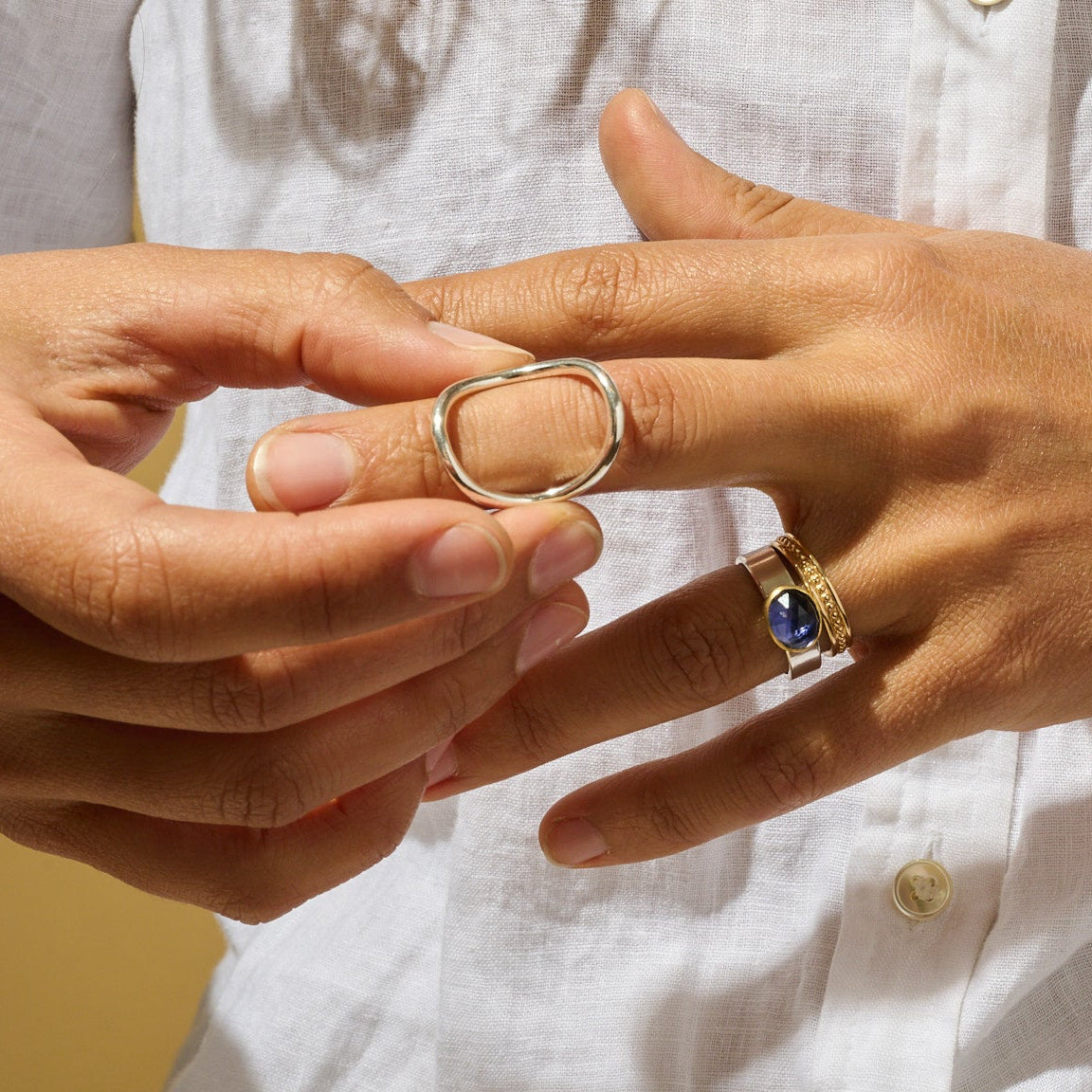 Close-up of two hands wearing rings, one with a silver circle ring and the other with a blue gemstone ring, stacked with a golds band against a warm background.