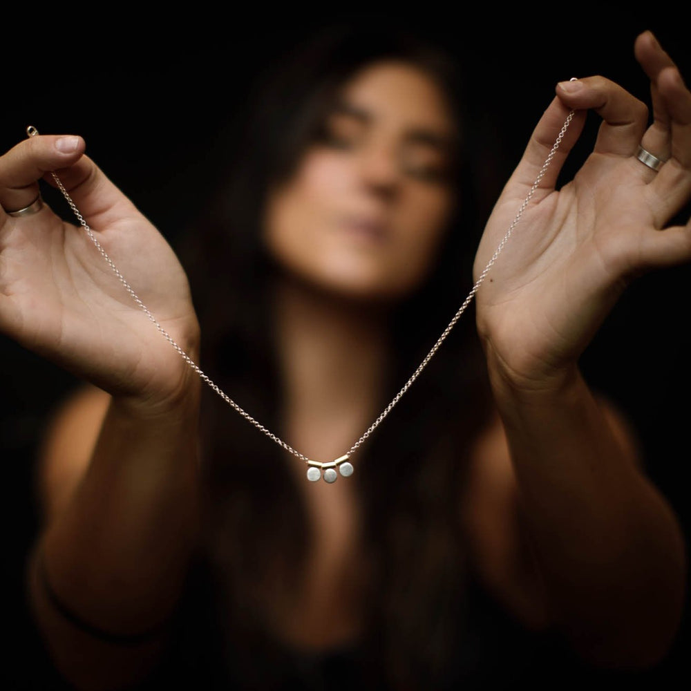 
                  
                    Holding a dainty, feminine necklace with three silver circle charms that dangle from yellow brass tubing hung on a silver chain.  
                  
                