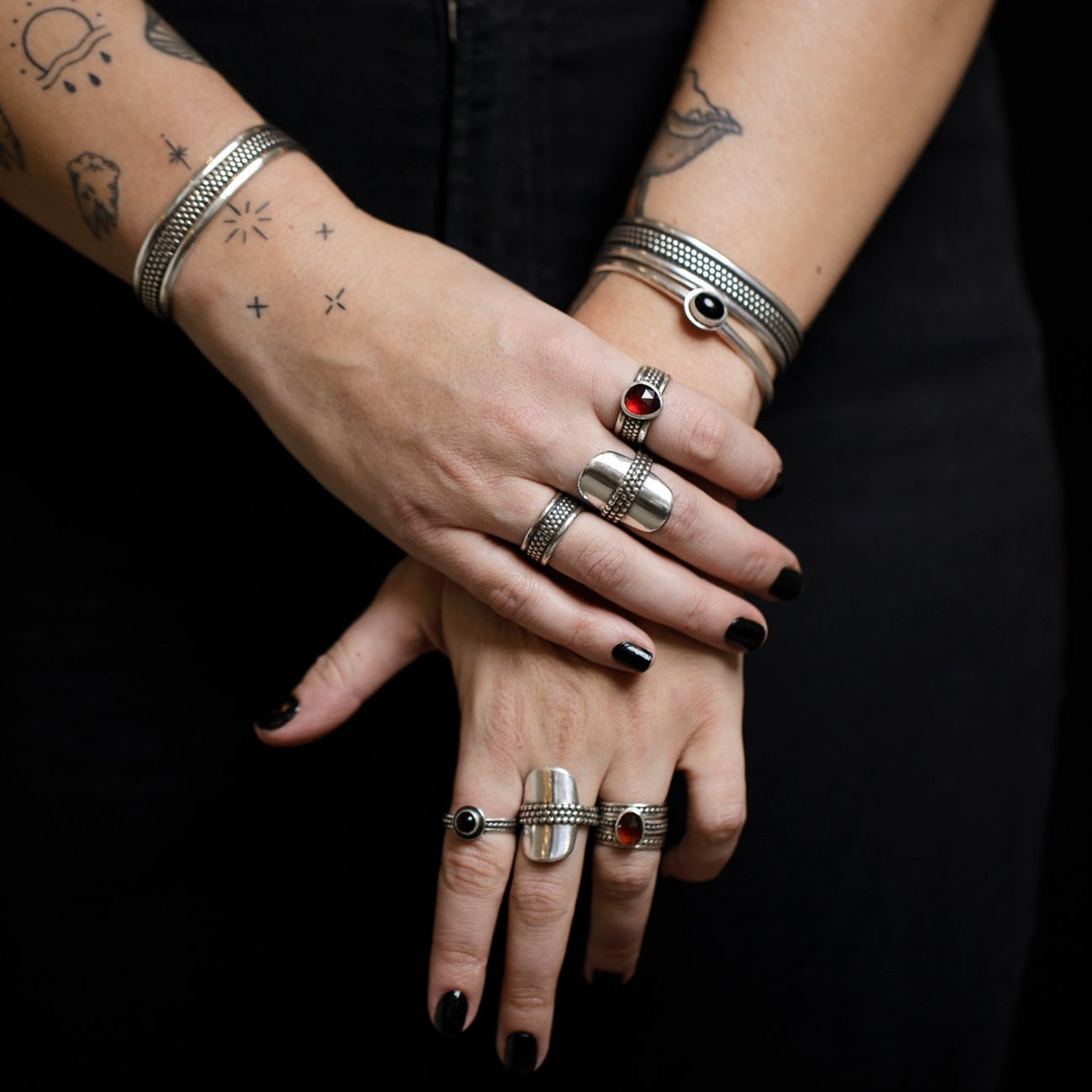 A woman standing with gently crossed hands wearing an assortment of hefty silver textured rings and bracelets against a dark background. 
