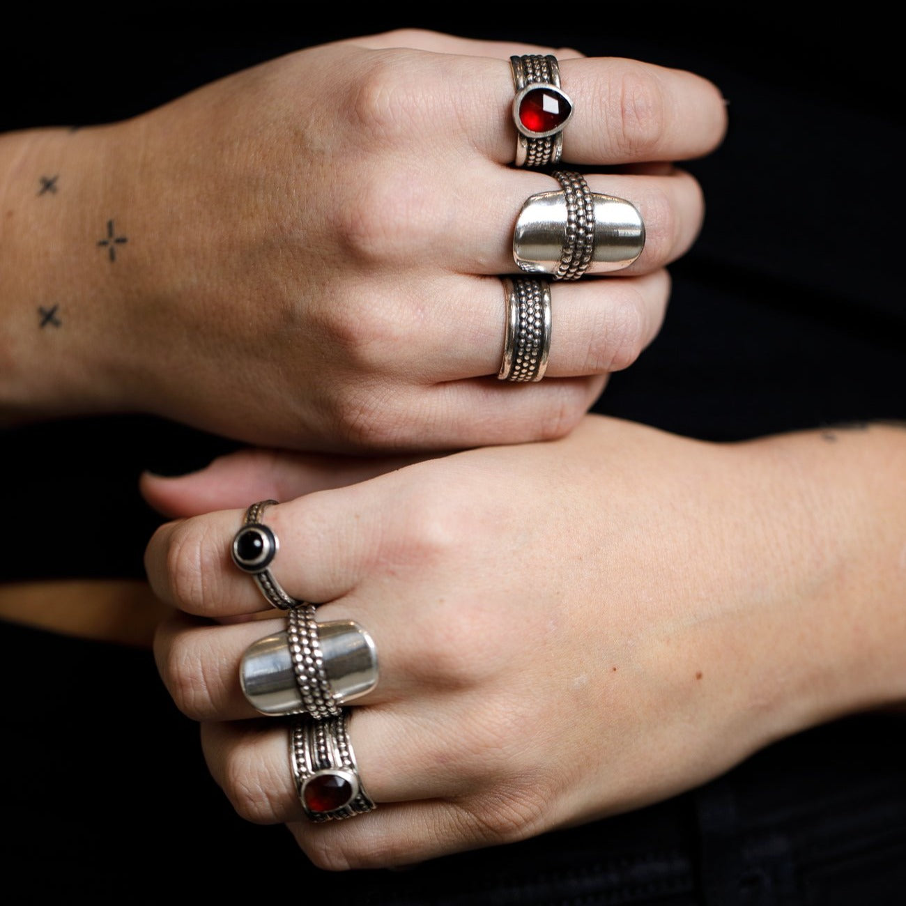 Two hands stacked together wearing an assortment of silver ring against a black background. 